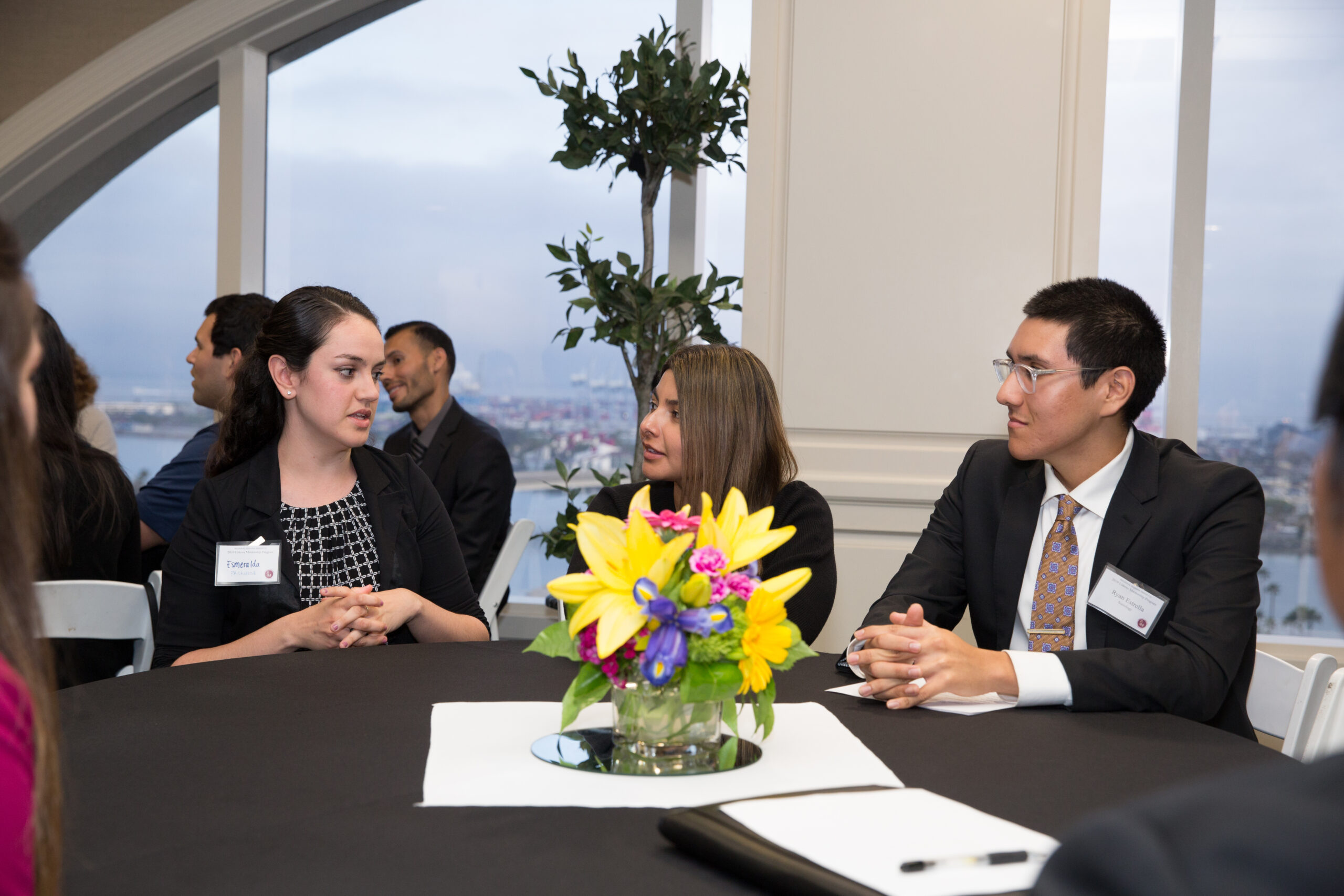Three professionals engage in a business discussion around a table with a floral centerpiece.